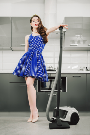 Beautiful young housewife wearing in a blue dress with white dots in the kitchen. Pin-up style. Fashion home shot. Full length portrait.の写真素材