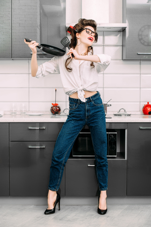 Beautiful girl is posing in everyday clothes with curlers in her hair and playing with kitchenware. Fashion home shot. Full length portrait.の写真素材