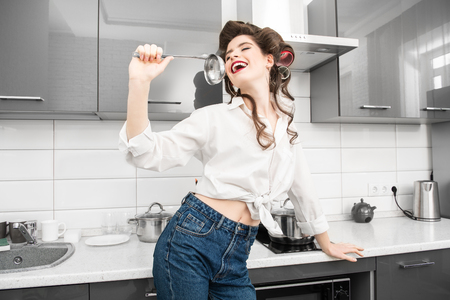 Beautiful girl is posing in everyday clothes with curlers in her hair and cooking. Fashion home shot. Pin-up style.の写真素材