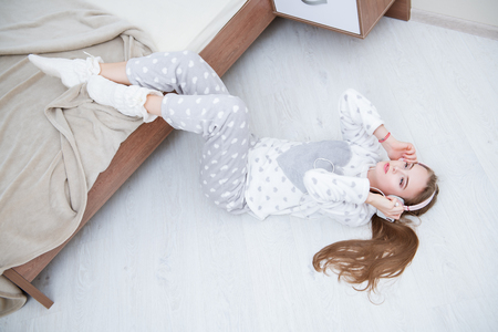 Portrait of an attractive young girl lying in a pajamas with headphones and smartphone on the floor near the bed at home. Beauty, cosmetics.の写真素材