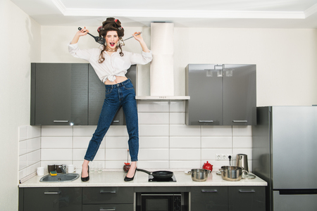 An attractive girl in everyday clothes and curlers stands on the countertop of the kitchen set and cooking. Fashion home shot. Full length portrait.の写真素材
