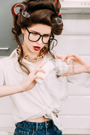 An attractive girl in a white shirt with curlers in her hair rubs a transparent glass to Shine. Pin-up style. Fashion home shot.の写真素材