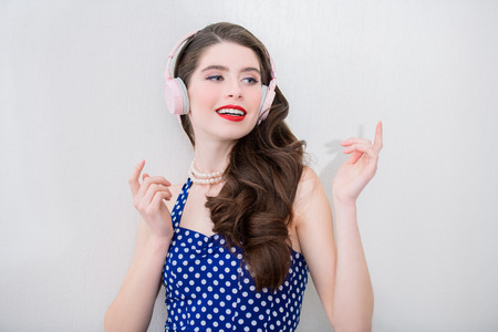 Portrait of attractive young woman in a blue dress with white dots with headphones on the white background of the wall. Pin-up style.の写真素材