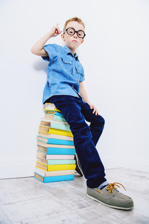 A portrait of a cute young boy wearing glasses sitting on the books and posing over the white background. Beauty, fashion, school, education.の写真素材