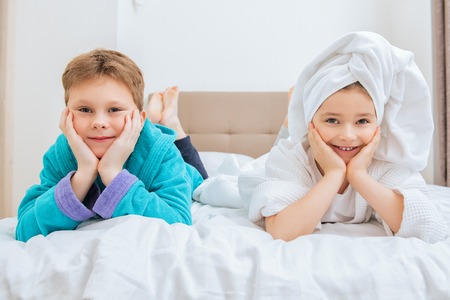 Cute girl and boy lie in bathrobes in bed at home. Fashion home shot. Childhood. Kid's fashion.の写真素材