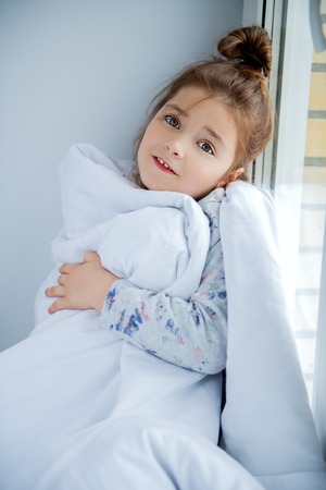 A little girl sittiing on a windowsill on a pillow covered with a blanket. Childhood. Kid's fashion.の写真素材