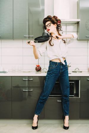 An attractive girl in everyday clothes and curlers stands in the kitchen set with a frying pan. Fashion home shot. Full length portrait.の写真素材
