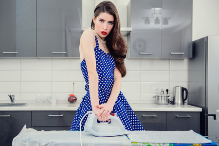 A portrait of an attractive young woman in a blue dress with white dots ironing clothes in the kitchen. Pin-up style.の写真素材