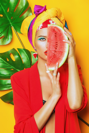 Fashionable woman with bright make-up eating watermelon. Yellow background. Beauty, fashion, make-up concept.の写真素材