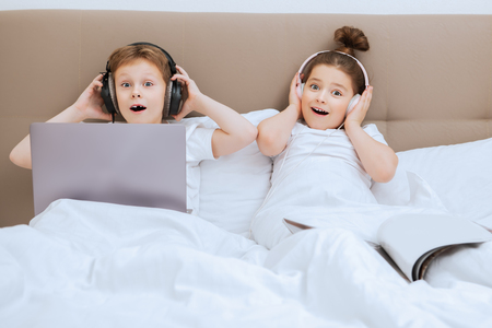 Surprised girl and boy lie in bed with laptop and magazine under the blanket in bed at home. Childhood. Kid's fashion.の写真素材
