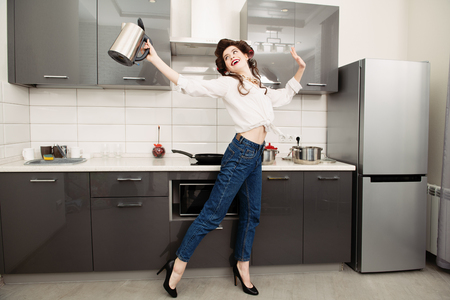 An attractive girl in everyday clothes and curlers stands in the kitchen with a kettle. Fashion home shot. Full length portrait.の写真素材