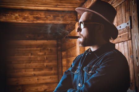 A portrait of a handsome man standing in front of the wooden house and smoking a cigar. Beauty, casual fashion.の写真素材
