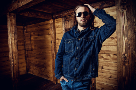A portrait of a handsome man standing in front of the wooden house. Beauty, casual denim fashion.の写真素材