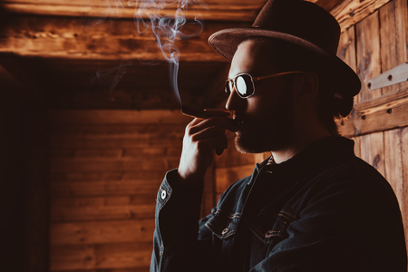 A portrait of a handsome man standing in front of the wooden house and smoking a cigar. Beauty, casual fashion.の写真素材
