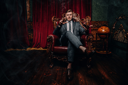 A full length portrait of a handsome mature man in a formal costume sitting in the armchair in the classic interior. Men's beauty, fashion.の写真素材