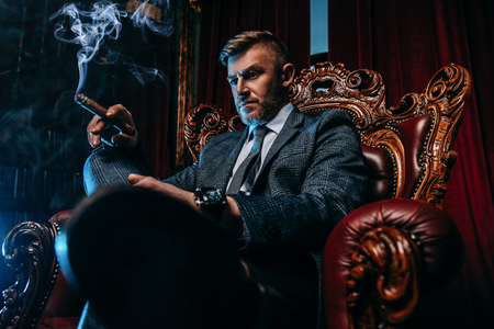 A portrait of a handsome mature man in a formal costume smoking a cigar in the armchair. Men's beauty, fashion.の写真素材