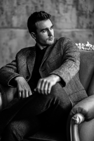 A portrait of a thoughtful fashionable young man sitting in a chair in the studio over the grunge background. Casual fashion for men.の写真素材