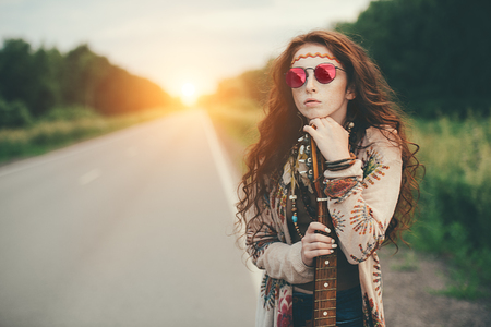 Beautiful hippie girl is standing with guitar on roadside outdoor. Contemporary bohemian style. Spirit of freedom. Bohemian, bo-ho style.の写真素材