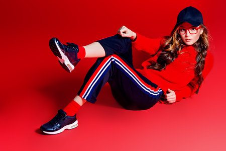 Teen style. Beautiful female model with long hair posing at studio in youth clothes. Red background....