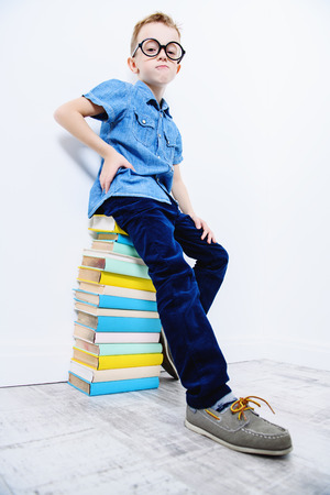 A portrait of a cute young boy wearing glasses sitting on the books and posing over the white background. Beauty, fashion, school, education.の写真素材