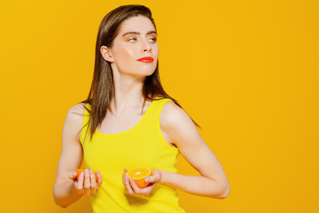 A portrait of a bright beautiful girl with a juicy orange posing in the studio over the yellow background. Beauty, health.の写真素材