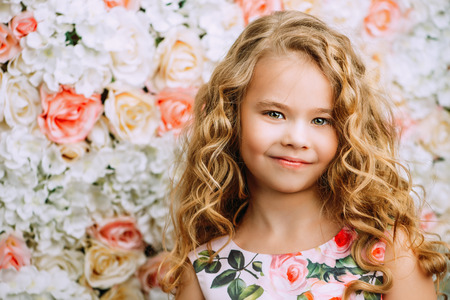 Portrait of a cute little girl in a beautiful summer dress with blonde curly hair. Background of roses. Kid's fashion, style.の写真素材