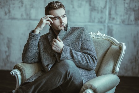 A portrait of a thoughtful fashionable young man sitting in a chair in the studio over the grunge background. Casual fashion for men.の写真素材