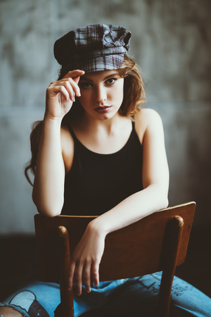 A portrait of a young beautiful woman in a cap sitting on the chair in the studio. Casual fashion, beauty.の写真素材