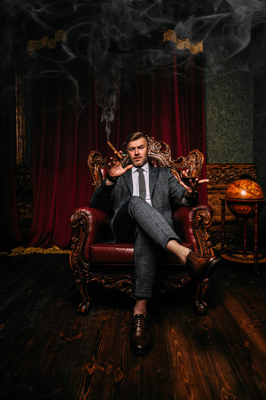 A full length portrait of a handsome mature man in a formal costume with a glass of wine and cigar sitting in the armchair in the classic interior. Men's beauty, fashion.の写真素材