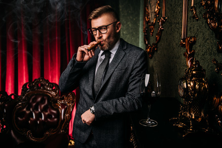 A portrait of a handsome mature man in a formal costume smoking a cigar in the interior. Men's beauty, fashion.の写真素材