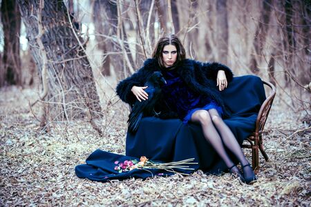A full length portrait of a beautiful mysterious girl in black posing on the bench in the forest. Beauty, fashion, nature.の写真素材