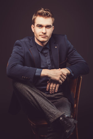 A portrait of a fashionable young man posing in black clothes sitting on a chair in the studio over...
