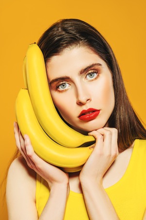 A close up portrait of a bright beautiful girl with a banana posing in the studio over the yellow background. Beauty, health.の写真素材