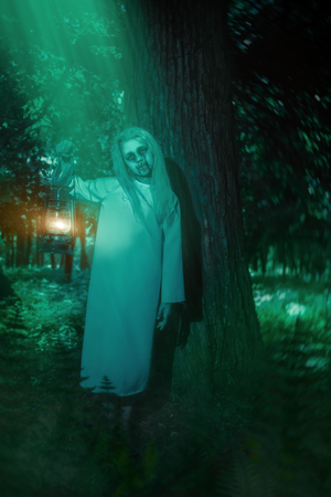 A full length portrait of a scary pale girl from a horror film in the forest with a lantern. Zombie, halloween.の写真素材