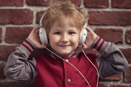 A portrait of a young schoolboy posing in the studio over the brick wall and listening to music in the headphones. Kids casual fashion, education.の写真素材
