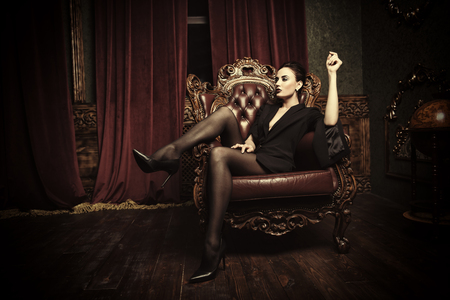 A full length portrait of a beautiful sexy woman wearing a black blazer and posing in classic interior on the armchair. Fashion, style, beauty, interior.の写真素材