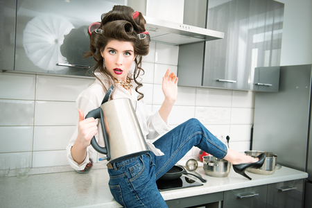 Beautiful girl is posing in everyday clothes with curlers in her hair and holding a kettle. Fashion home shot. Pin-up style.の写真素材