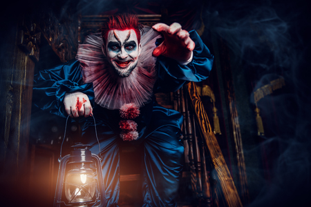 A portrait of an angry crazy clown from a horror film with a lantern on the stairs. Halloween, carnival.の写真素材