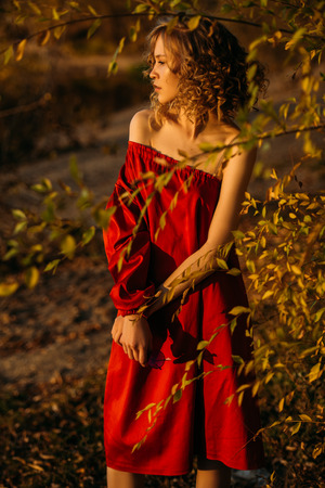A beautiful young girl is posing in the countryside in a red dress. Fashion concept. Golden autumn.の写真素材