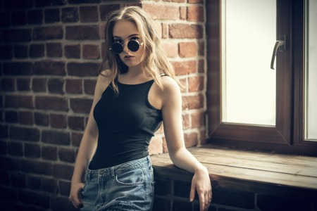 A portrait of a young beautiful woman in sunglasses posing in the studio over the brick wall near a window. Casual fashion, beauty, optics.の写真素材