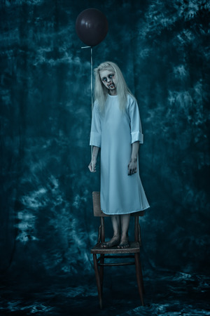 A full length portrait of a scary pale girl from a horror film in the forest with a baloon on the chair. Zombie, halloween.の写真素材