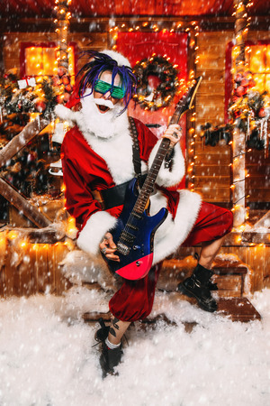 Cool rocker in Santa Claus costume and luminous glasses plays the guitar near a festively decorated house.の写真素材