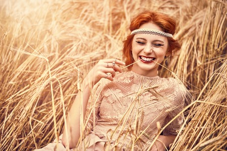 Romantic young woman with beautiful red hair lies on a wheat field and laughs. Beauty, fashion. Modern hippie, bohemian style.の写真素材