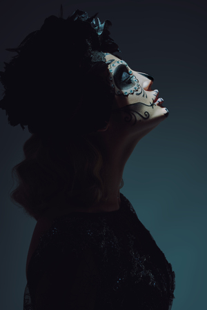 Side view portrait of Calavera Catrina in the dark. A ray of light falls on her face. Young woman with sugar skull makeup. Dia de los muertos. Day of The Dead. Halloween.の写真素材