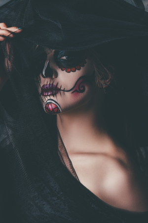 Portrait of a beautiful and scary Calavera Catrina in black dress and black hat with a veil over dark background. Sugar skull makeup. Dia de los muertos. Day of The Dead. Halloween.の写真素材