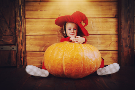 Funny little girl in a witch costume sits behind a big pumpkin over wooden background. Halloween.の写真素材