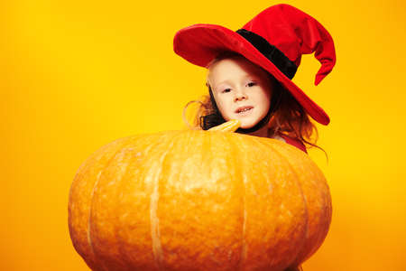 Halloween. Funny little girl in a witch costume poses with a big pumpkin over yellow background. Copy space.の写真素材