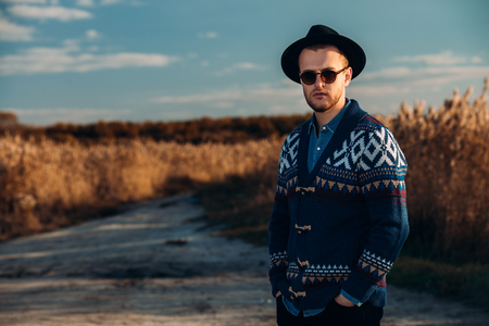 Autumn fashion. Portrait of a handsome man wearing cardigan and hat standing outdoor over autumn nature background.の写真素材
