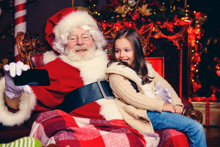 Santa Claus takes a selfie sitting on a chair with a cute little girl at home. Merry Christmas and Happy New Year.の写真素材