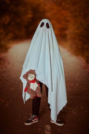 Lonely ghost of a child under a white sheet stands with his teddy bear in a dark forest. Halloween.の写真素材